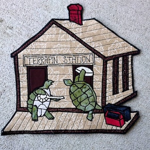 Puede incluir: Una casa acolchada marrón y beige con una chimenea roja y las palabras "TERRAPIN STATION" sobre la puerta. Dos tortugas verdes están de pie en un porche de madera frente a la casa. Una tortuga sostiene un cartel que dice "TERRAPIN STATION".