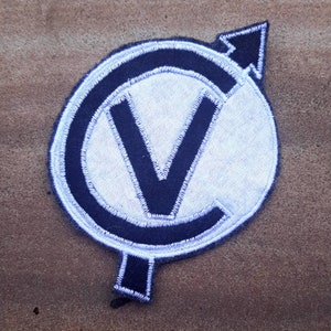 May include: A black and white embroidered patch with a white circle and a black letter V inside. The circle has a black outline and a white outline. An arrow points to the right.