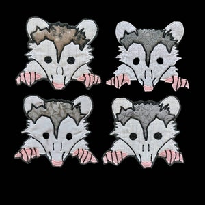 May include: Four white fabric patches with embroidered opossum faces. Each opossum has a different pattern on its back, including a brown and white speckled pattern, a gray and white swirl pattern, a gray and white speckled pattern, and a gray and white swirl pattern.