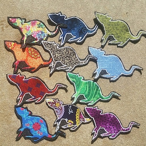 May include: Ten colourful rat-shaped fabric patches with different patterns. The patches are all the same size and shape, and they are arranged in a circle on a light brown surface.