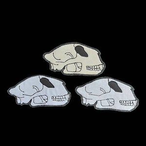 May include: Three embroidered patches of animal skulls in different shades of gray. The skulls are all facing the right side of the image.