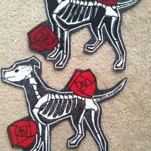 May include: Two black and white embroidered patches of skeleton dogs with red roses. The patches are on a gray fabric background.