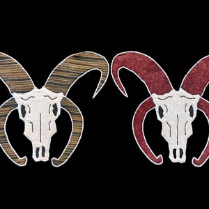 May include: Two embroidered ram skulls, one with brown and gold stripes and the other with red stripes, on a black background.