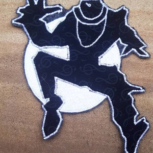 May include: Black and white patch featuring a stylized figure in a dynamic pose. The figure is outlined in white stitching and filled with a black fabric patterned with musical notes. The figure is set against a white circle.