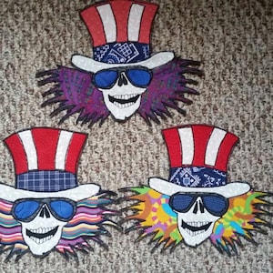 Puede incluir: Tres parches bordados con un cráneo que lleva un sombrero de copa con rayas rojas, blancas y azules y un pañuelo. Cada parche tiene un fondo de color diferente con un diseño psicodélico.