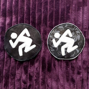 Op de afbeelding: Twee zwarte en witte geborduurde patches met een gestileerd stick figure-ontwerp. De patches zijn rond en hebben een zwarte achtergrond met een witte stick figure. Een patch heeft een bloemenpatroon op de achtergrond.