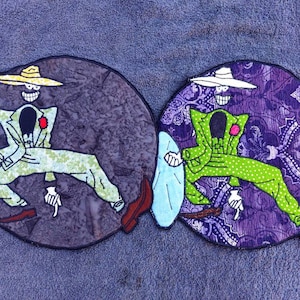 Puede incluir: Dos parches bordados con un personaje de dibujos animados con un traje verde y un sombrero, sosteniendo un platillo. Un parche tiene un fondo morado con un patrón floral, el otro tiene un fondo gris.