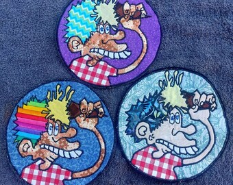 May include: Three embroidered patches featuring cartoon characters with colorful hair, wearing red and white checkered shirts, and holding a brown object in their right hand. The patches are circular and have different backgrounds: purple, blue, and green.