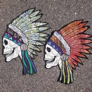 Op de afbeelding: Twee kleurrijke geborduurde patches met Indiaanse hoofdtooien met schedelgezichten. De hoofdtooien zijn gemaakt van stof met ingewikkelde patronen en kleuren. Een hoofdtooi is blauw, groen en paars, en de andere is rood, oranje en geel.