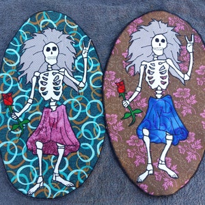 May include: Two embroidered patches featuring skeletons with peace signs and roses. One patch has a blue background with a pink skirt and the other has a brown background with a blue skirt.