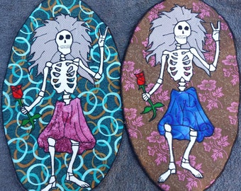 May include: Two embroidered patches featuring skeletons with peace signs and roses. One patch has a blue background with a pink skirt and the other has a brown background with a blue skirt.