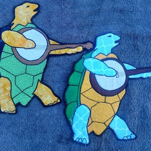 Puede incluir: Dos figuras de tortugas bordadas de colores tocando el banjo. Las tortugas son verdes, azules, amarillas y naranjas. Los banjos son marrones con cuerdas blancas.