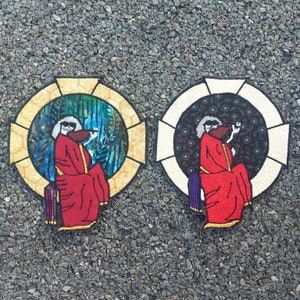 Puede incluir: Dos parches bordados que representan una figura con una túnica roja y una cara de calavera. Los parches son circulares y tienen un borde dorado y blanco. Un parche tiene un fondo verde y azul, mientras que el otro tiene un fondo negro.