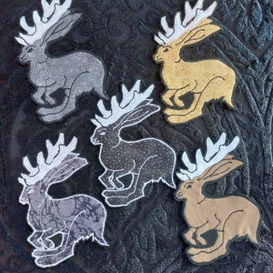 Op de afbeelding: Vijf geborduurde patches van hazen met geweien, elk in een andere kleur en patroon. De patches bevinden zich op een zwarte stoffen achtergrond.