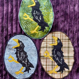 Puede incluir: Tres aplicaciones de pájaros negros con picos y patas amarillas. Cada aplicación está sobre un fondo de tela diferente: verde con un patrón de hojas, azul con un patrón floral y marrón con un patrón de cuadros.