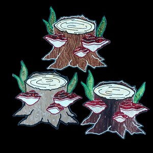 Könnte beinhalten: Drei gestickte Aufnäher, die Baumstümpfe mit darauf wachsenden roten Pilzen darstellen. Die Baumstümpfe sind braun mit einem hellbraunen Holzmaserungsmuster. Die Pilze haben weiße Unterseiten und rote Oberseiten.