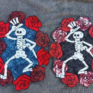 May include: Two embroidered patches featuring a skeleton figure wearing a top hat and dancing among red roses. The patches are on a gray background.