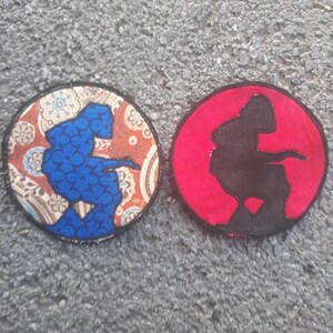 May include: Two round embroidered patches, one with a blue silhouette of a person on a paisley patterned background and the other with a black silhouette of a person on a red background.