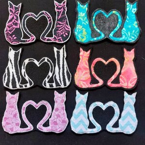 Op de afbeelding: Negen paar stoffen kattenapplicaties in verschillende kleuren en patronen, elk paar vormt een hartvorm met hun staarten.