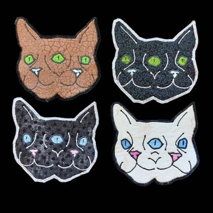 May include: Four embroidered patches featuring three-eyed cats. The cats are brown, black, white, and black with white spots. Each cat has three green or blue eyes and a white outline.
