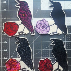 Puede incluir: Cuatro aplicaciones de tela de cuervos posados ​​sobre rosas. Los cuervos son negros, negros con un patrón floral y dos tonos de gris diferentes. Las rosas son rojas, rosas y moradas.