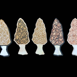 Puede incluir: Cinco parches bordados de morillas en diferentes tonos de marrón y beige. Las setas están delineadas con hilo negro y tienen un tallo blanco.