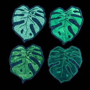 May include: Four green monstera leaf shaped patches with a dark green outline. The patches are made of a textured fabric.