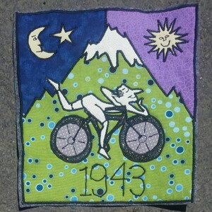 Pode incluir: Um remendo de tecido verde, azul e roxo com uma cena de montanha branca. Uma pessoa est&aacute; a andar de bicicleta para cima da montanha. O ano de 1943 est&aacute; bordado no centro inferior do remendo.