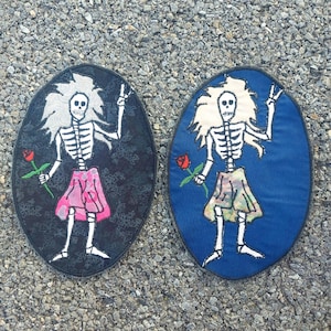 May include: Two embroidered patches featuring skeleton figures with flowers. One patch is on a black background with a pink skirt and the other is on a blue background with a multi-colored skirt.