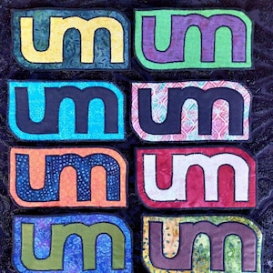May include: Eight fabric patches with the letter "M" in different colors and patterns. The patches are arranged in two rows of four. The colors include yellow, green, blue, black, orange, red, purple, and brown.
