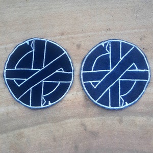 Puede incluir: Dos parches bordados en blanco y negro con un diseño circular. El diseño es un círculo con una cruz en su interior, la cruz está hecha de una línea blanca gruesa. El círculo está bordeado de blanco.