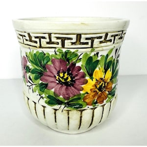 May include: A white ceramic planter with a hand-painted floral design. The flowers are in shades of purple, yellow, and orange, with green leaves. The planter has a Greek key pattern around the top and vertical lines at the bottom.