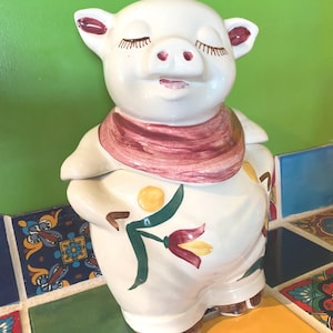 May include: A white ceramic pig-shaped cookie jar with a red scarf and floral accents. The jar has closed eyes and a smiling expression. The jar is decorated with green leaves and red flowers.