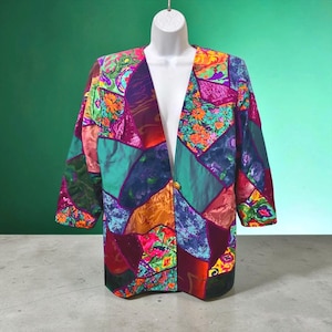 May include: A long-sleeve blazer featuring a vibrant patchwork design with various geometric shapes in shades of magenta, teal, orange, purple, and blue. The garment has a single gold-toned button closure at the center and a collarless neckline.