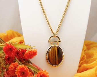 May include: A gold-toned necklace with a large oval pendant featuring a tiger's eye stone. The pendant is framed in gold and hangs from a textured chain. Orange flowers and fabric are in the background.