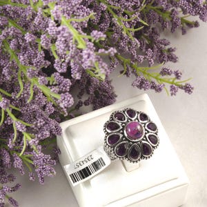 May include: A silver-toned ring with a flower-shaped design, featuring a central pink stone surrounded by dark purple gemstones. The ring is displayed on a white surface, with a barcode tag and a backdrop of purple flowers.