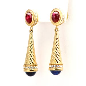 May include: A pair of gold-toned earrings with a red gemstone at the top, surrounded by small clear stones. The earrings have a long, cone-shaped design with a spiral texture and a blue gemstone at the bottom.