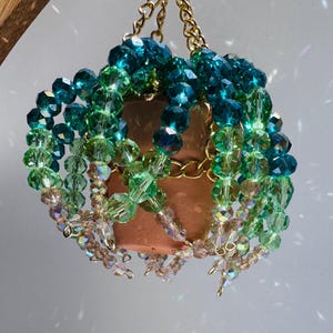 May include: A hanging planter with a copper-colored pot, adorned with strands of green and blue crystal beads. The planter is suspended by a gold chain and hook, creating a decorative and eye-catching piece.