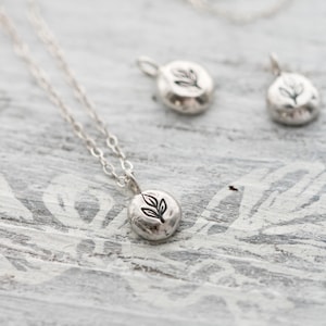 Puede incluir: Un collar de plata con un peque&ntilde;o colgante redondo con un dise&ntilde;o de hoja. El colgante est&aacute; en una cadena de plata delicada.