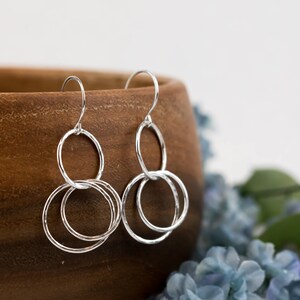 May include: A pair of silver dangle earrings with three interlocking circles. The earrings are hanging from a wooden bowl.