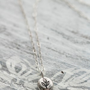 Puede incluir: Un collar de plata con un peque&ntilde;o colgante redondo con un dise&ntilde;o de hoja. El colgante est&aacute; en una cadena delicada.