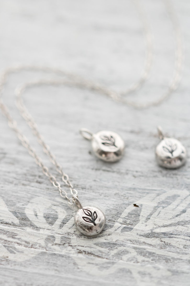 Puede incluir: Un collar de plata con un peque&ntilde;o colgante redondo con un dise&ntilde;o de hoja. El colgante est&aacute; en una cadena de plata delicada.