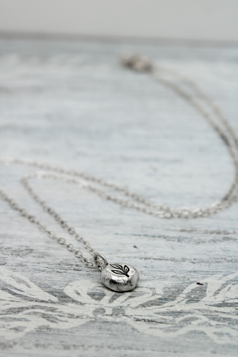 Puede incluir: Un collar de plata con un peque&ntilde;o colgante redondo con un dise&ntilde;o de hoja. El colgante tiene una superficie texturizada.
