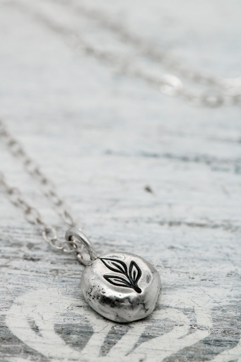 Puede incluir: Un collar de plata con un peque&ntilde;o colgante redondo con un dise&ntilde;o de hoja. El colgante tiene una superficie texturizada y est&aacute; unido a una cadena de plata delicada.