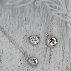 Puede incluir: Tres collares colgantes de plata con un dise&ntilde;o de hoja. Los colgantes son de forma ovalada y tienen una superficie texturizada. Los collares est&aacute;n sobre una superficie de madera blanca.