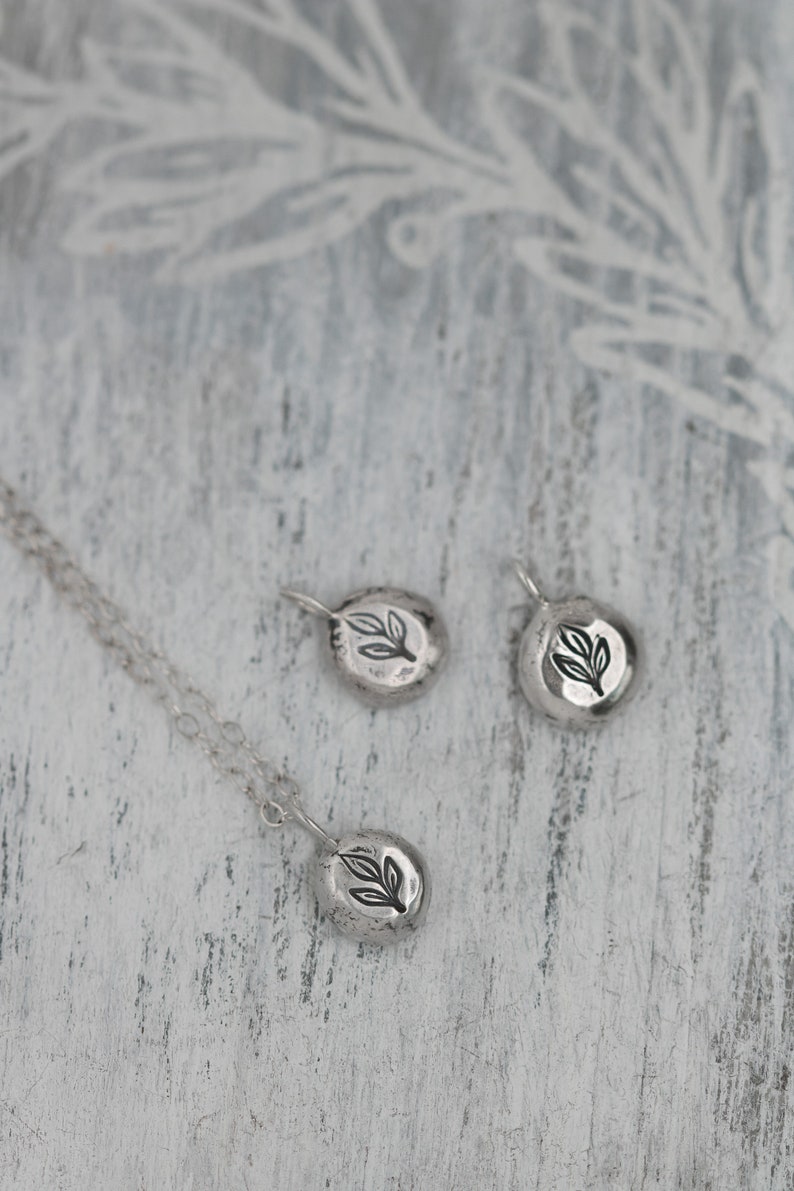Puede incluir: Tres collares colgantes de plata con un dise&ntilde;o de hoja. Los colgantes son de forma ovalada y tienen una superficie texturizada. Los collares est&aacute;n sobre una superficie de madera blanca.