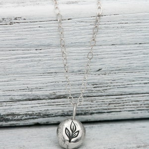 Puede incluir: Un collar de plata con un peque&ntilde;o colgante redondo que presenta un dise&ntilde;o de hoja estilizado. El colgante tiene una superficie texturizada y la cadena es de estilo delicado y enlazado.
