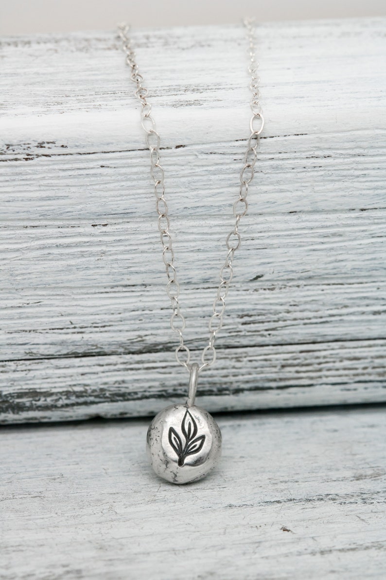 Puede incluir: Un collar de plata con un peque&ntilde;o colgante redondo que presenta un dise&ntilde;o de hoja estilizado. El colgante tiene una superficie texturizada y la cadena es de estilo delicado y enlazado.