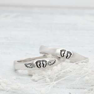 May include: Two silver rings with engraved baby footprints. One ring features a pair of footprints, while the other ring has footprints with angel wings.