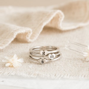 May include: Three silver rings stacked together, each with a heart-shaped charm. The charms are engraved with the letters "g", "m", and "s". The rings are displayed on a beige linen surface, with small white flowers and a hair pin.